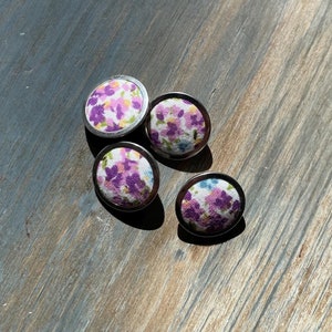 Op de afbeelding: Vier ronde stoffen knopen met een zilveren metalen rand. De knopen hebben een witte achtergrond met een bloemenpatroon in tinten paars en geel.