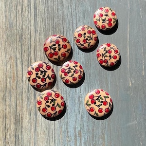 Pode incluir: Uma coleção de oito botões redondos de madeira com um design de estampa de cerejas. Os botões são bege com cerejas vermelhas e hastes verdes.