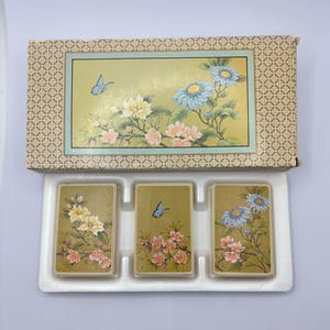 May include: A rectangular box with a beige geometric pattern and a central illustration of blue flowers, pink blossoms, and a blue butterfly. Below the box, three decks of cards are arranged in a white plastic tray, each featuring floral designs.