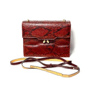 May include: A rectangular shoulder bag featuring a deep red and brown snakeskin pattern with a textured finish. The bag has a structured flap closure with gold-toned metal hardware and a thin, adjustable matching strap with a light tan underside.