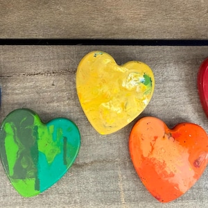 May include: Five heart-shaped wax crayons arranged in a row. The colors include a dark blue, a bright green, a yellow, an orange, and a red heart. Each heart has a smooth, slightly marbled texture and a rounded, solid form made of melted wax.