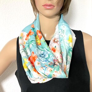 Op de afbeelding: Een lichtblauwe infinity-sjaal met een levendige bloemenprint in oranje, gele en witte tinten. De stof toont een aquarelachtig patroon van diverse bloemen en groen blad, gedrapeerd in een cirkelvormige lus rondom de hals van de drager.