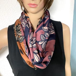 Puede incluir: Una bufanda infinita con un estampado botánico de hojas estilizadas en tonos naranja óxido, azul marino oscuro y malva apagado. La tela tiene un acabado suave y satinado, dispuesta en un bucle circular alrededor del cuello.