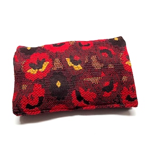 May include: A rectangular, woven clutch with a floral pattern. The dominant color is a deep red, with accents of black, yellow, and gold. The clutch is likely made of a textured fabric, and is set against a white background.