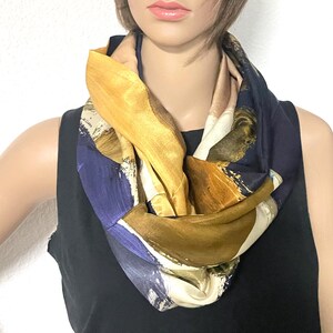 Op de afbeelding: Een infinity sjaal met een gedurfde abstracte print met vegende penseelstreken in tinten diep marineblauw, metallic goud, oker en crème. De stof heeft een gladde, glanzende afwerking en is gedrapeerd in een cirkelvormige lus rond de nek.