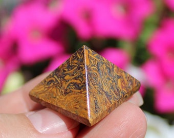 May include: A small, brown and gold pyramid-shaped crystal with intricate patterns. The crystal is being held in a hand against a blurred background of pink and white flowers.