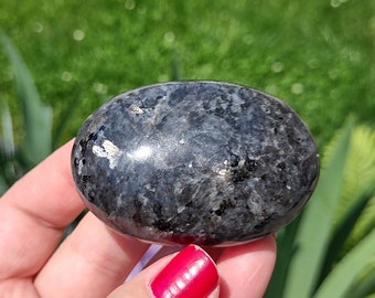 May include: A smooth, oval-shaped black gemstone with silver flecks. The stone is being held in a hand.