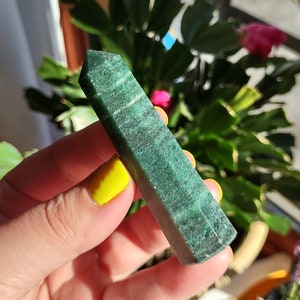 Puede incluir: Una punta de cristal verde oscuro con una superficie lisa y pulida. El cristal se sostiene en una mano con esmalte de uñas amarillo.