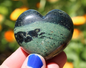 Puede incluir: Una piedra en forma de corazón verde y negro con un patrón de remolino. La piedra está siendo sostenida en una mano con esmalte de uñas azul.