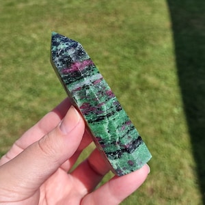 May include: A green, black, and pink zultanite crystal point, also known as a zoisite, is held in a hand. The crystal is smooth and polished, with a pointed tip.