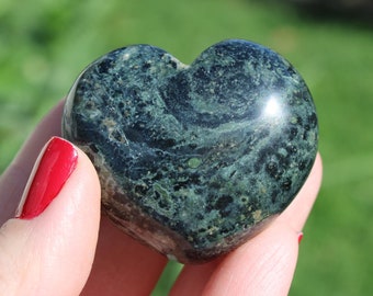 Puede incluir: Una piedra en forma de corazón con un patrón veteado verde oscuro y negro. La superficie pulida refleja la luz, y la piedra se sostiene en una mano con esmalte de uñas rojo. El fondo es verde borroso.