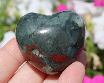 Puede incluir: Una piedra de jaspe verde y roja en forma de corazón con un acabado pulido. La piedra se sostiene en una mano sobre un fondo de flores blancas y rosas.