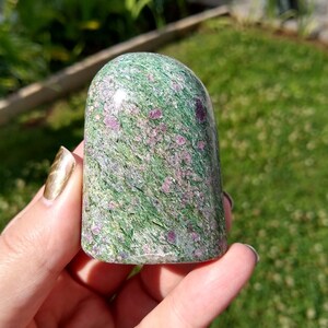 May include: A green and pink ruby zoisite crystal, also known as ruby in zoisite, in a polished, rounded shape. The crystal has a smooth, glossy finish and is speckled with pink ruby inclusions.