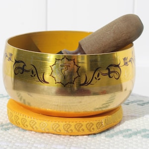 May include: A gold-coloured singing bowl with a wooden mallet sits on a yellow cushion. The bowl has black decorative patterns and a central symbol. The cushion is embroidered with a repeating design. The bowl is used for meditation.