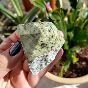 May include: A green and gray crystal with black flecks, shaped like a pyramid, is held in a hand. The crystal is about 2 inches tall.