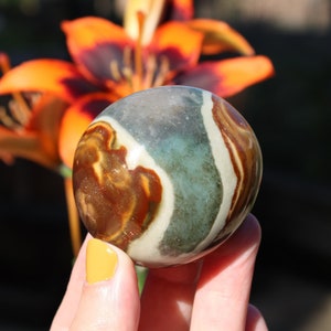 Può includere: Una sfera di pietra rotonda, liscia, a strisce verdi, marroni e bianche. La pietra è tenuta in una mano.