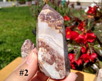 May include: A white, brown, and pink banded agate crystal point with a pointed tip. The crystal is approximately 10 cm tall and is held in a hand. The background is a blurred image of a house and a garden.