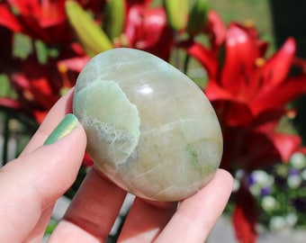 May include: A smooth, green and white polished stone with a rounded shape. The stone is being held in a hand.
