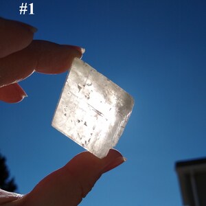 May include: A clear, translucent, square-shaped crystal with a slight shimmer. The crystal is being held up to the sun, and the light is shining through it.