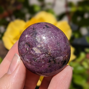 May include: A smooth, polished purple and black gemstone with a rounded shape. The gemstone is held in a hand.