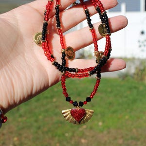 May include: A beaded necklace featuring alternating red and black glass beads with small gold-toned spacers. The necklace is accented with several small gold-toned coin charms and a central pendant shaped like a red glitter heart with gold wings.