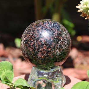 May include: A polished granite sphere with a speckled pattern of dark red and black, resting on a clear, faceted glass stand. The sphere is displayed outdoors with green foliage and a white flower in the background.