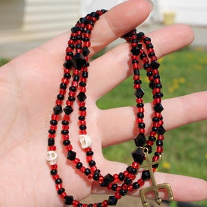 May include: A layered necklace with red and black beads, interspersed with black geometric shapes and small white skull beads. A gold-colored trident charm hangs from the necklace.