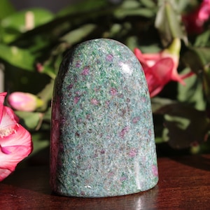 May include: A green and pink ruby in zoisite crystal, polished and shaped into a point, with a smooth, rounded top. The crystal is sitting on a wooden surface.