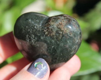 Puede incluir: Una piedra verde oscura en forma de corazón con marcas marrones y negras. La piedra es lisa y pulida.