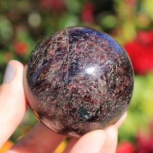 Könnte beinhalten: Eine polierte, dunkelrote Granat-Kugel mit einer glatten, glänzenden Oberfläche. Die Kugel ist aus Naturstein gefertigt und hat ein einzigartiges, komplexes Muster von Einschlüssen.