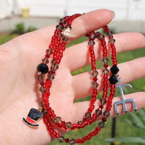 May include: A layered beaded necklace with red and brown beads. The necklace features charms including a top hat, a skull, and a silver trident. The beads are a mix of small round and faceted shapes.