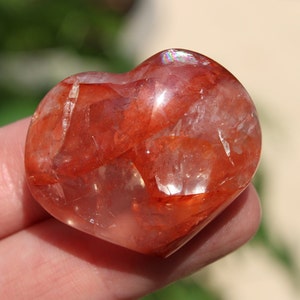 Puede incluir: Un cristal en forma de corazón con una base transparente y clara y inclusiones rojas, naranjas y amarillas. El cristal tiene un ligero brillo iridiscente.