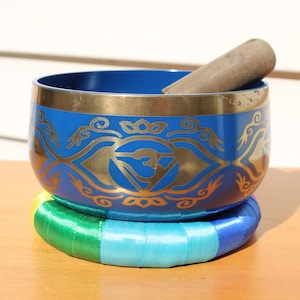 May include: A blue singing bowl with gold decorative patterns and a wooden striker. The bowl rests on a colourful, circular cushion with bands of green, yellow, blue, and turquoise. This item is used for meditation and sound healing.