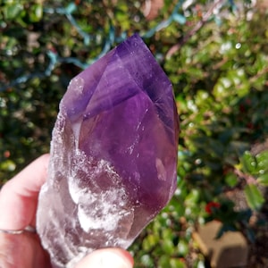 Könnte beinhalten: Ein großer, violetter Amethystkristall mit einer klaren, weißen Basis. Der Kristall ist oben spitz und hat eine natürliche, raue Textur.