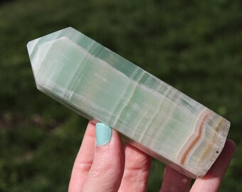 May include: A green calcite crystal obelisk with a polished, rectangular shape. The crystal has a layered appearance with bands of white and tan hues. The obelisk is held against a blurred green background.
