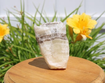 Pode incluir: Um cristal de calcita branco e cinza com bandas, com uma textura áspera e natural. O cristal está sentado sobre uma superfície de madeira.