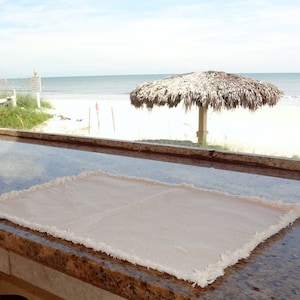 May include: A light beige table runner with frayed edges sits on a brown granite countertop. The runner is in focus, while the background shows a beach scene with a thatched roof umbrella.