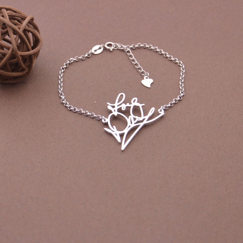 May include: A silver bracelet with a delicate chain and a charm that says "Love" in cursive script. The charm is shaped like a branch with a heart at the end.