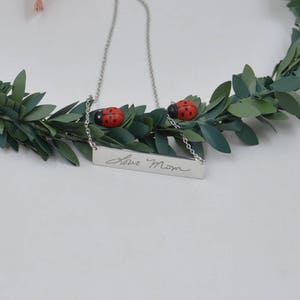 May include: Silver necklace with a rectangular pendant engraved with the handwritten message "Love Mom". The necklace is adorned with two small red ladybug charms.