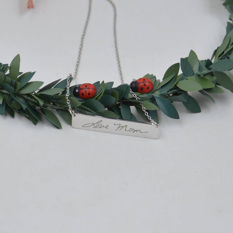 May include: Silver necklace with a rectangular pendant engraved with the handwritten message "Love Mom". The necklace is adorned with two small red ladybug charms.