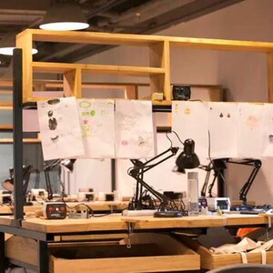 May include: A wooden workbench with multiple black adjustable desk lamps and white papers with drawings on them. The workbench has a wooden shelf above it.