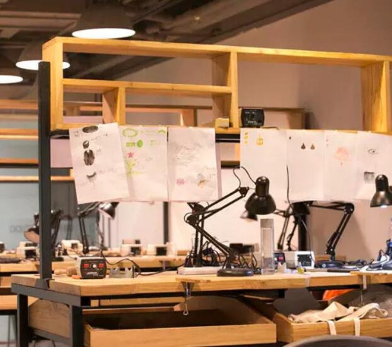 May include: A wooden workbench with multiple black adjustable desk lamps and white papers with drawings on them. The workbench has a wooden shelf above it.