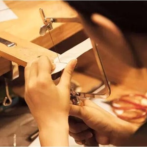 May include: A person uses a jeweler's saw to cut a piece of metal. The saw is held in a wooden frame and the metal is clamped in place. The person is wearing a white shirt and has their hands on the saw.