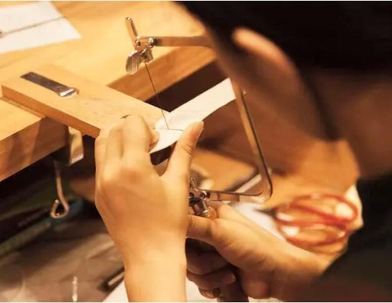 May include: A person uses a jeweler's saw to cut a piece of metal. The saw is held in a wooden frame and the metal is clamped in place. The person is wearing a white shirt and has their hands on the saw.
