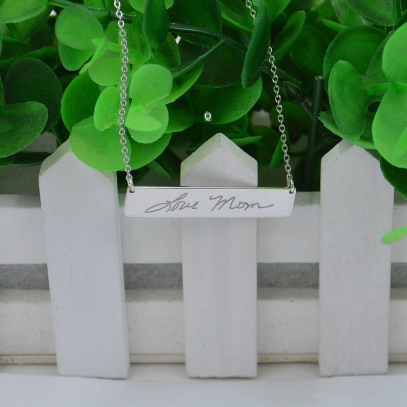May include: A silver necklace with a bar pendant that has the words "Love Mom" engraved on it. The necklace is hanging against a white picket fence with green foliage in the background.