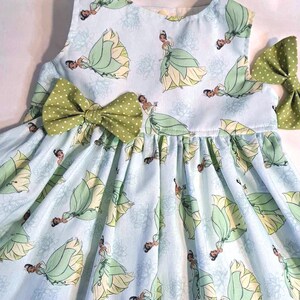 May include: A light blue sleeveless dress with a princess pattern and green bows. The dress features a repeating pattern of a princess in a green and yellow gown. Two green bows with white polka dots are attached to the dress.