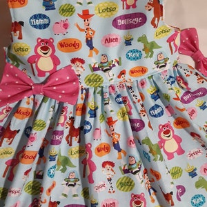 Puede incluir: Un vestido azul claro sin mangas con un lazo rosa. El vestido est&aacute; cubierto de un patr&oacute;n de personajes de Toy Story, incluyendo Woody, Buzz Lightyear, Jessie y Lotso. Los personajes est&aacute;n rodeados de burbujas de di&aacute;logo con sus nombres.