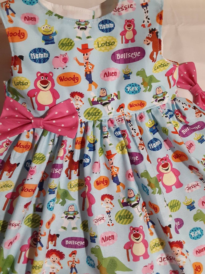 Puede incluir: Un vestido azul claro sin mangas con un lazo rosa. El vestido est&aacute; cubierto de un patr&oacute;n de personajes de Toy Story, incluyendo Woody, Buzz Lightyear, Jessie y Lotso. Los personajes est&aacute;n rodeados de burbujas de di&aacute;logo con sus nombres.