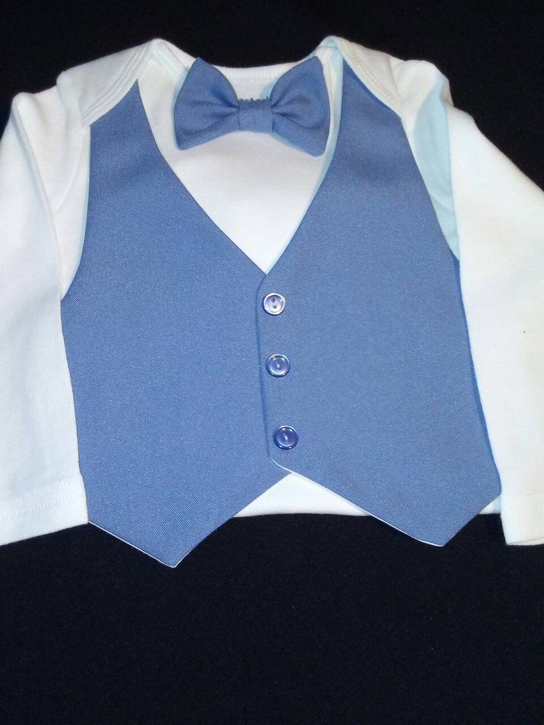 以下が含まれることがあります： 白いボディスーツに青いベストと青い蝶ネクタイをつけた赤ちゃん。ベストには5つのボタンがついています。