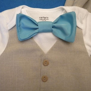 May include: A white bodysuit with a light blue bow tie and a beige linen vest with two brown buttons. The bodysuit has the text "carter's made with love" printed on the front.